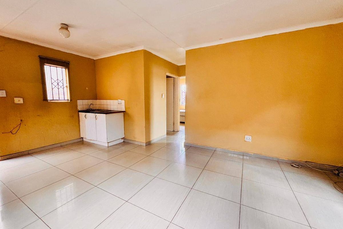 An empty, open-plan kitchen area featuring tiled flooring, basic base cabinetry with a sink, and security bars on the window.