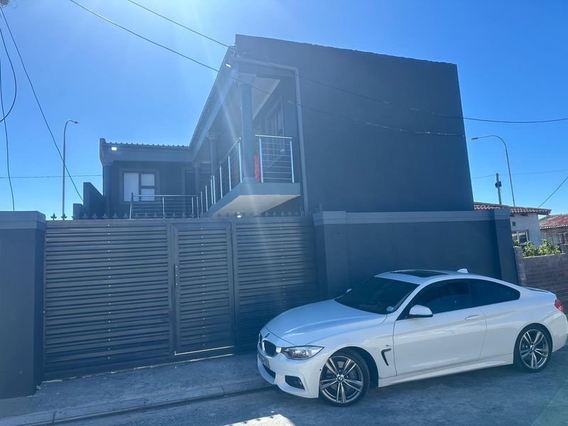 Front elevation of a modern two-story house featuring a dark exterior finish, a secure metal gate, and an upper-level balcony with stainless steel railings.