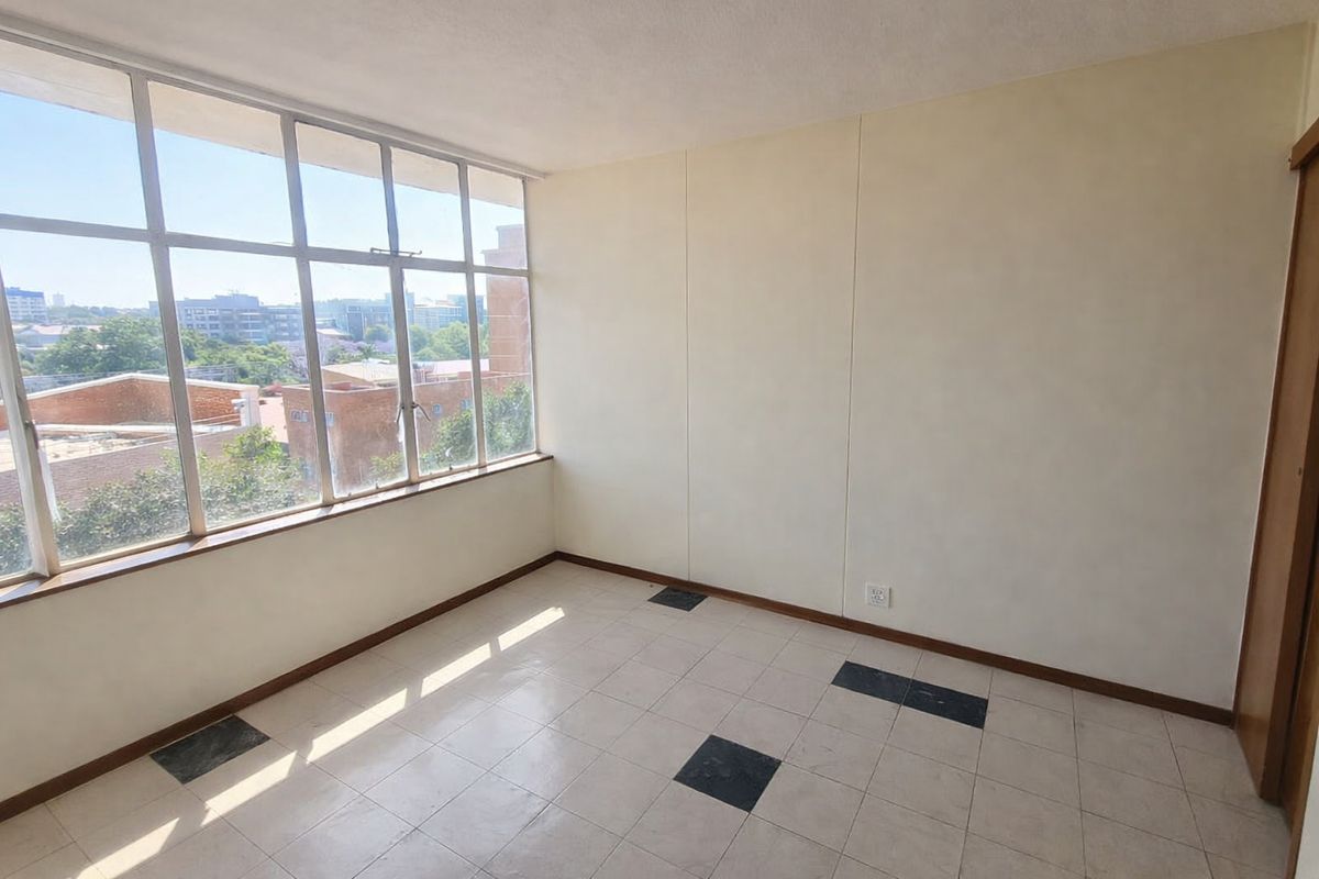 An empty, sunlit living area featuring large metal-framed windows with city views and tiled flooring with decorative accents.