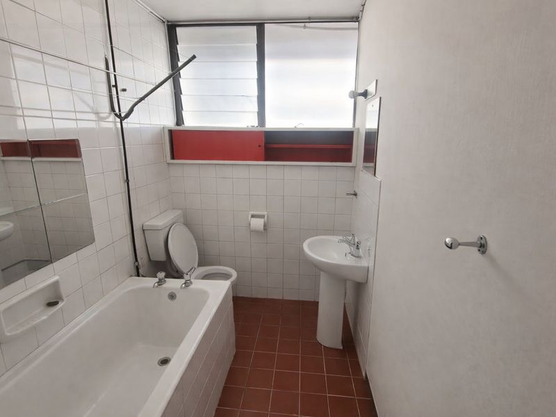 A functional bathroom featuring a bathtub, pedestal basin, toilet, and tiled finishes, with louvre windows providing natural ventilation.