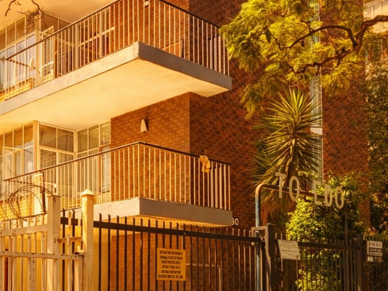 Street-level view of a multi-story brick apartment building featuring private balconies with metal railings and a secure gated entrance.