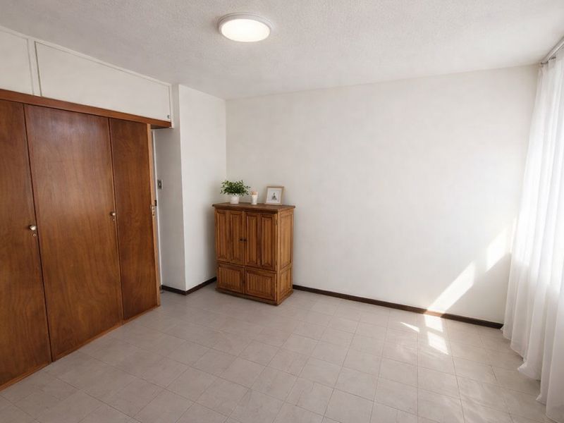 A bright, neutral bedroom featuring built-in wooden cupboards and tiled flooring, offering a clean and versatile space ready for personalization.