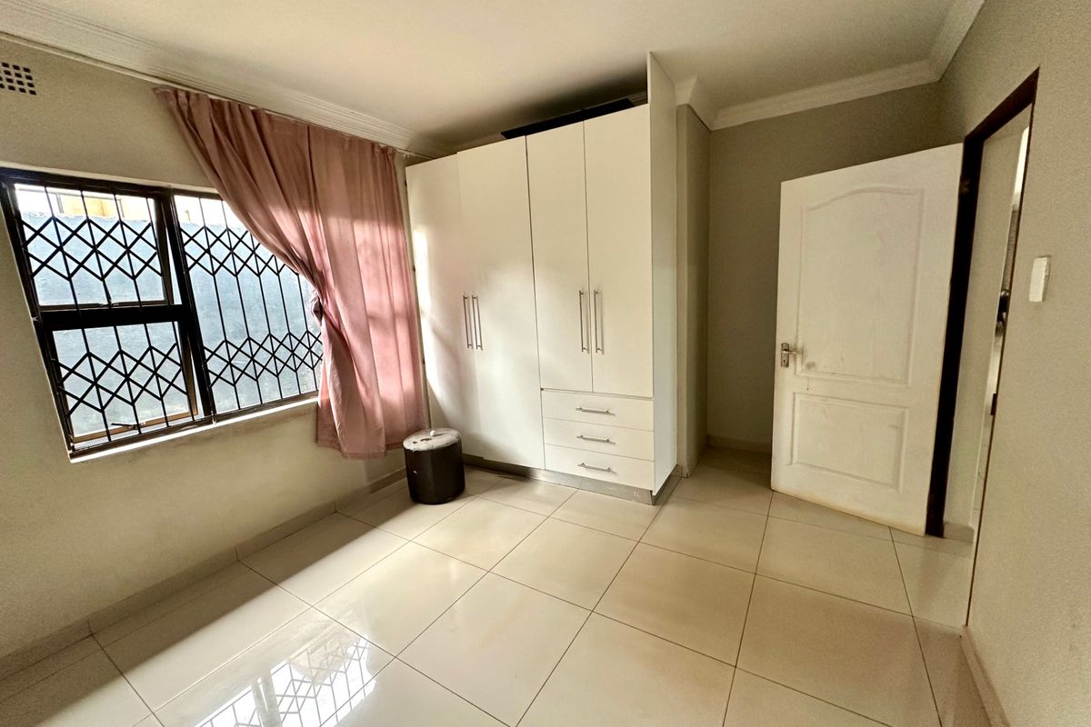A bright, empty bedroom featuring tiled flooring, a built-in wardrobe, and a window fitted with a security gate.