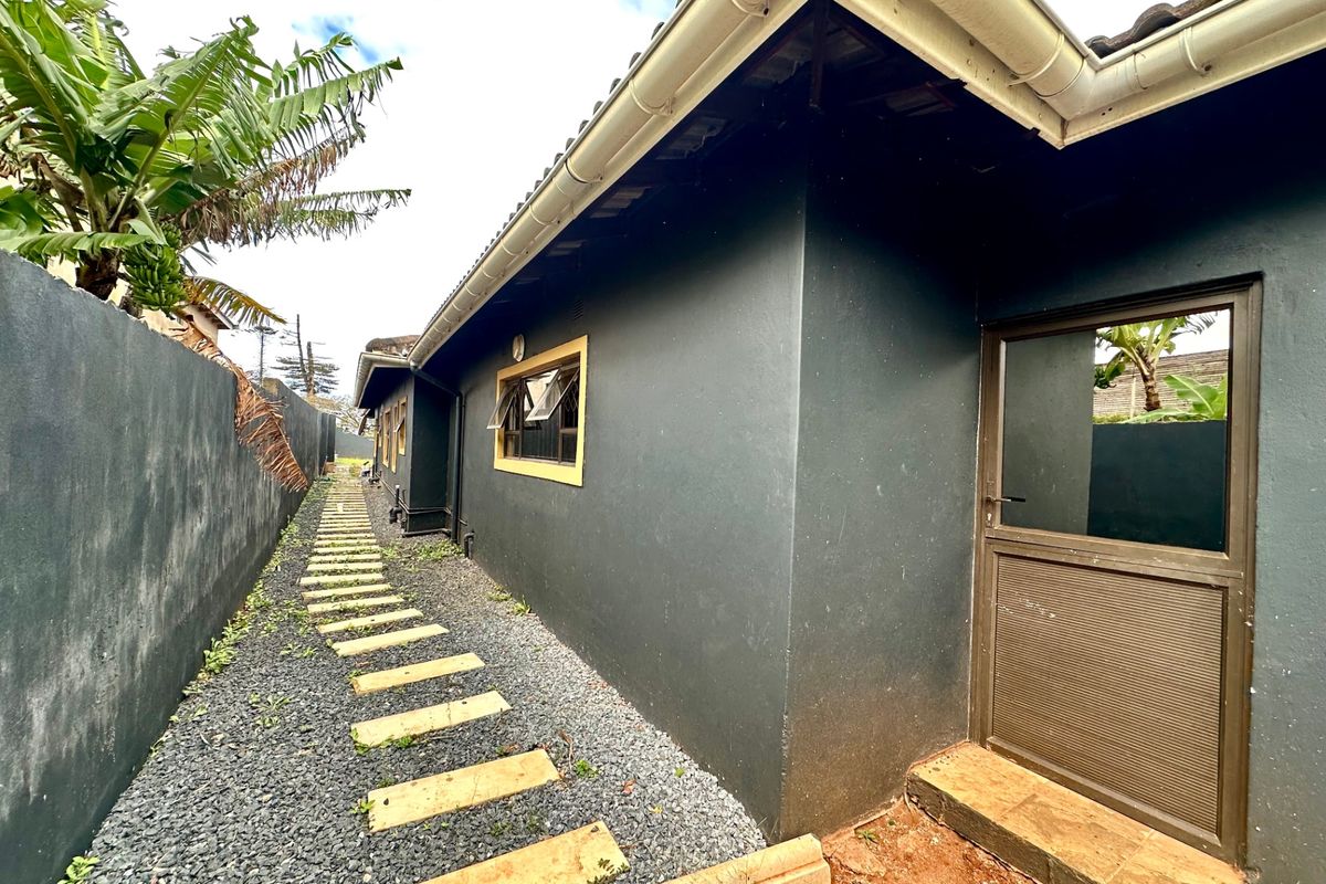 Side exterior view of the property featuring a dark painted facade, aluminium door and window frames, and a gravel pathway with stone pavers.