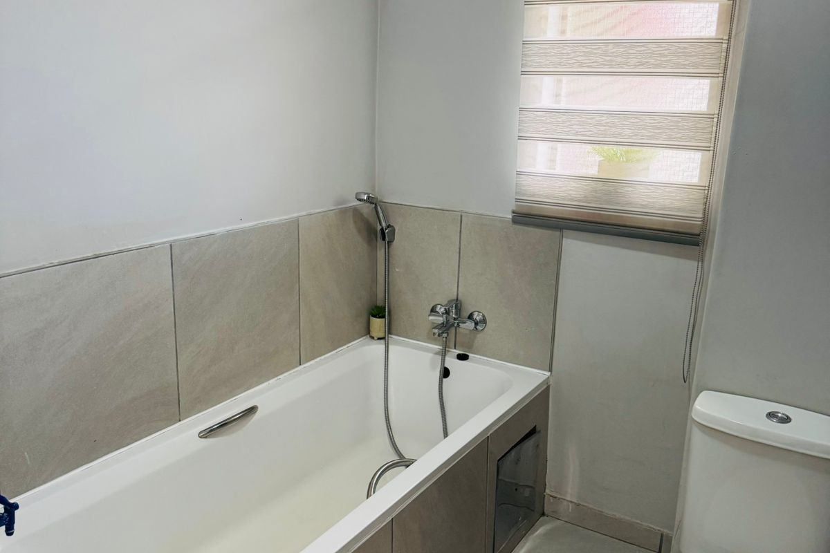 A clean and functional bathroom featuring a built-in bathtub with a handheld shower mixer, a standard toilet, and neutral-toned floor and wall tiling.