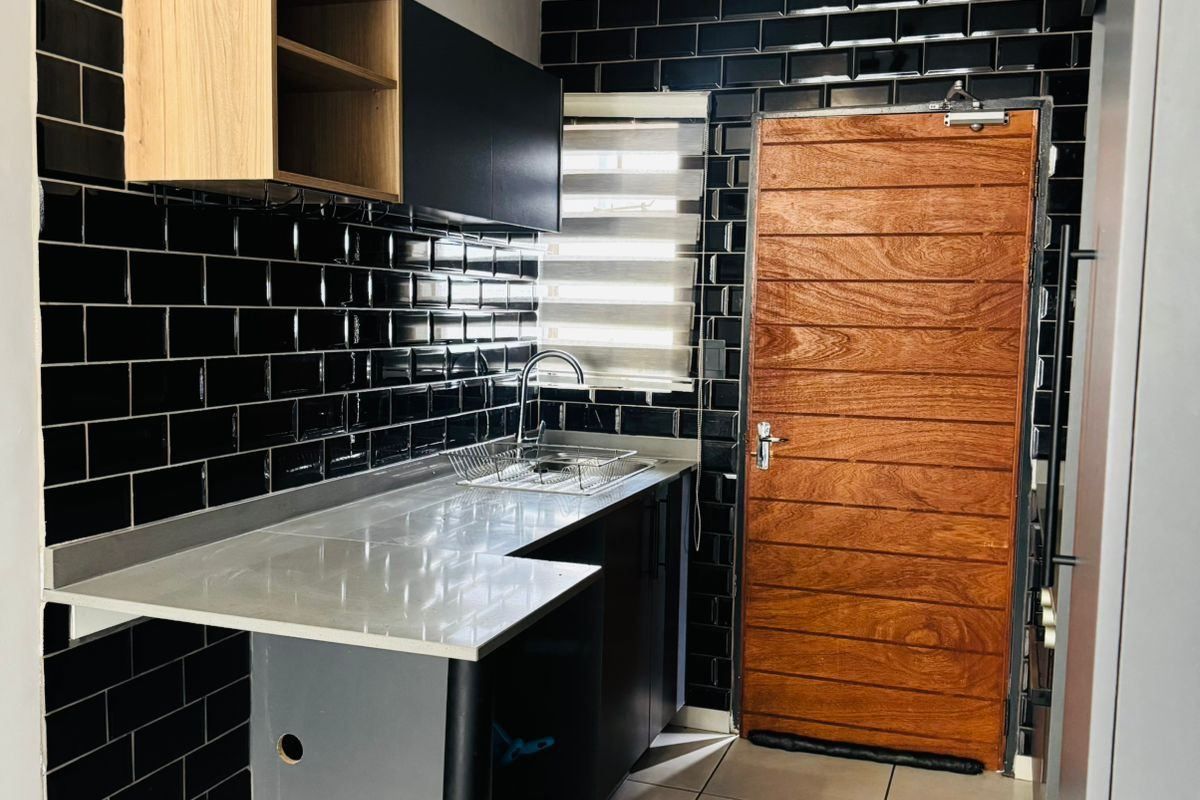 Modern kitchen featuring black subway tile backsplash, stainless steel countertop with integrated sink, and sleek cabinetry, complemented by a timber door and tiled flooring.