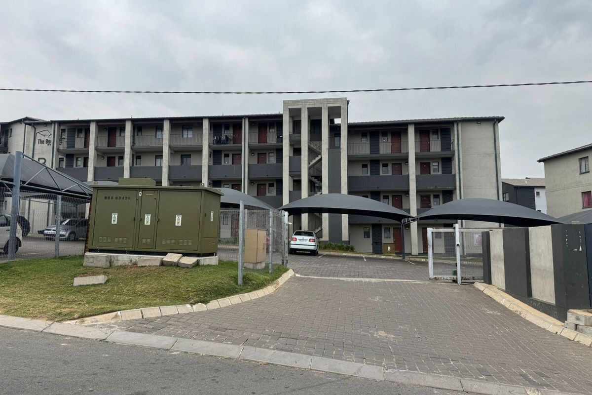 Exterior view of a multi-story apartment complex featuring paved parking areas, shade cloth carports, and a central concrete stairwell.