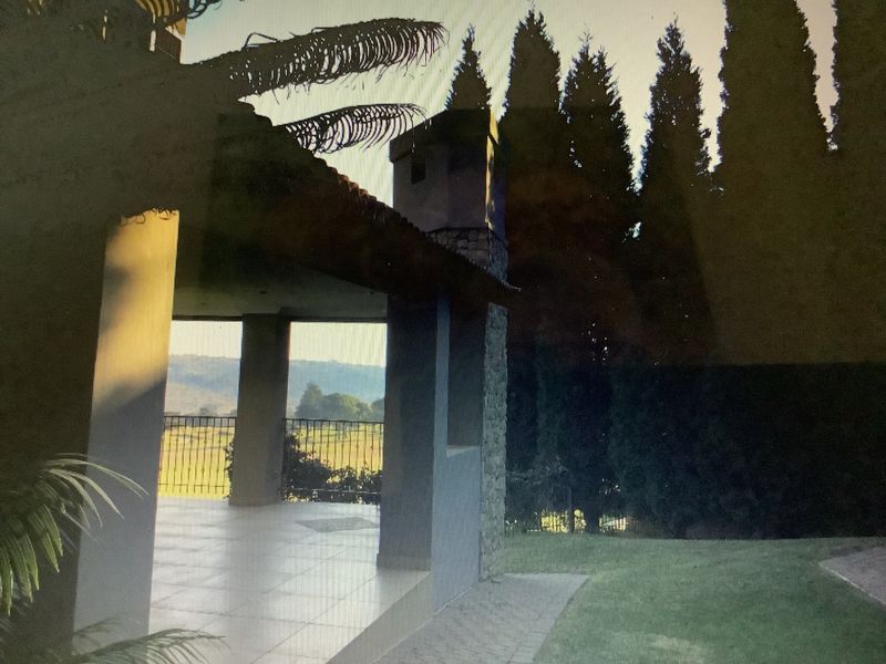 A covered outdoor patio area featuring a stone-clad chimney and metal balustrade, overlooking a garden and distant landscape.