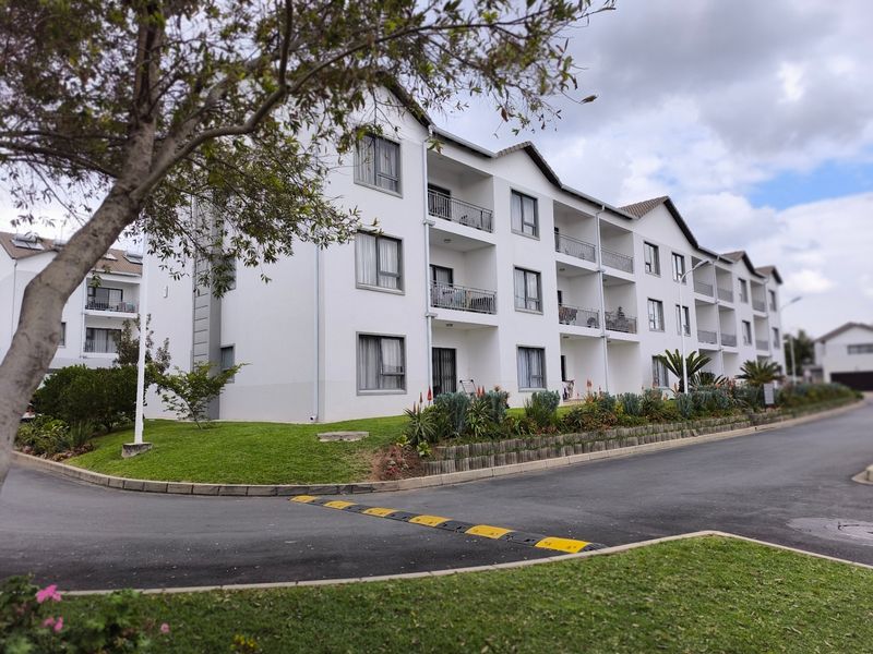A well-maintained multi-story apartment complex featuring private balconies, landscaped garden beds, and a paved driveway with speed control measures.