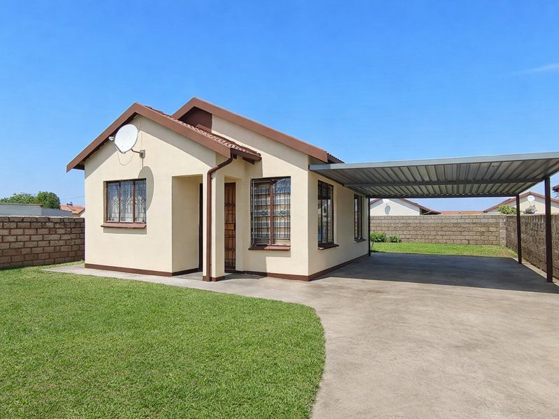 A well-maintained single-story house featuring a paved driveway, attached carport, and a spacious front lawn enclosed by a perimeter wall.