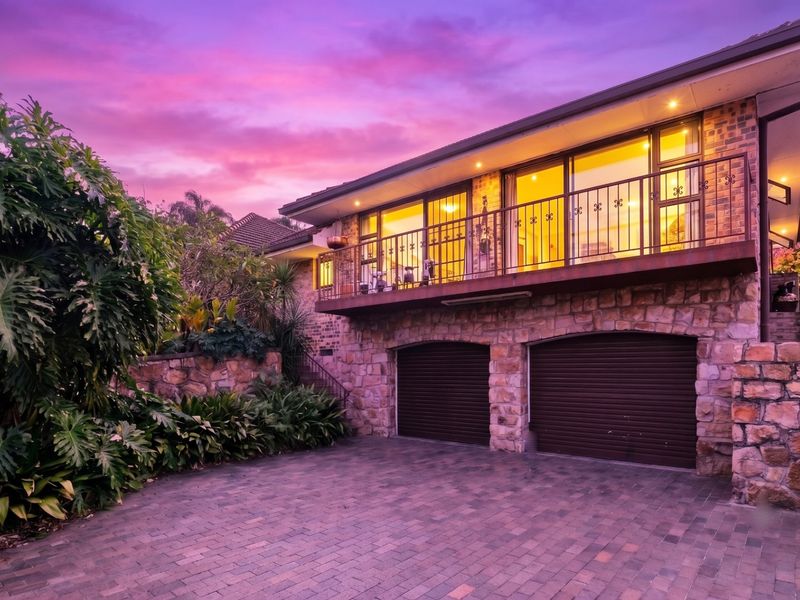 Front elevation featuring a double garage with roller doors, stone masonry facade, and an upper-level balcony with wrought iron railings.