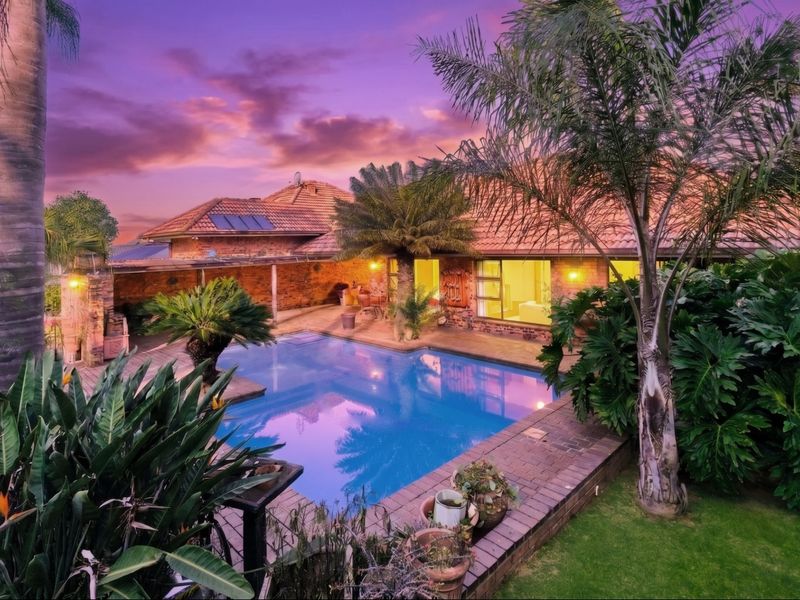A well-maintained outdoor pool area featuring a swimming pool, brick paving, and a covered patio, set against a sunset backdrop.