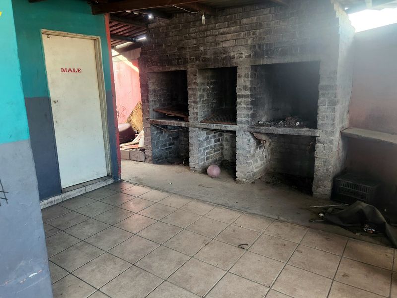 Covered outdoor patio area featuring a large built-in brick braai, tiled flooring, and exposed timber roof structure, requiring maintenance and cosmetic updates.