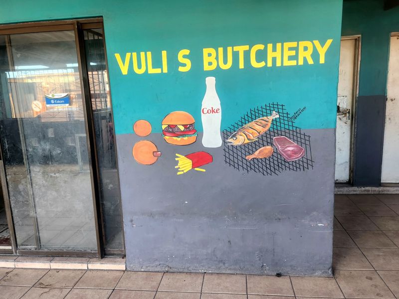 Exterior view of a commercial butchery storefront featuring a painted mural on the facade, tiled flooring, and a sliding glass entrance door.