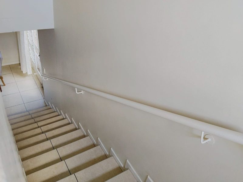 A clean, minimalist staircase featuring neutral-toned tiled steps and a simple white wall-mounted handrail, leading down to a tiled landing area.