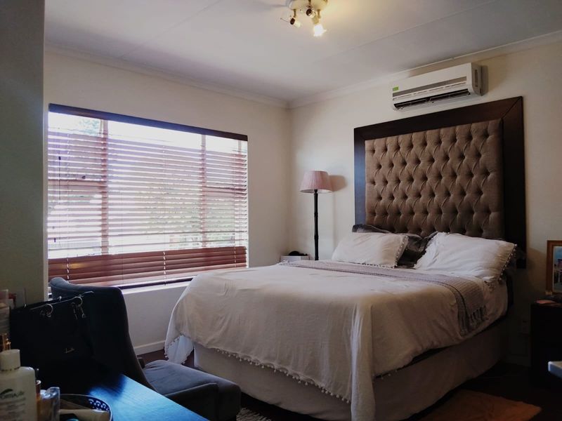A well-maintained bedroom featuring a wall-mounted air conditioning unit, large window with venetian blinds, and neutral finishes.