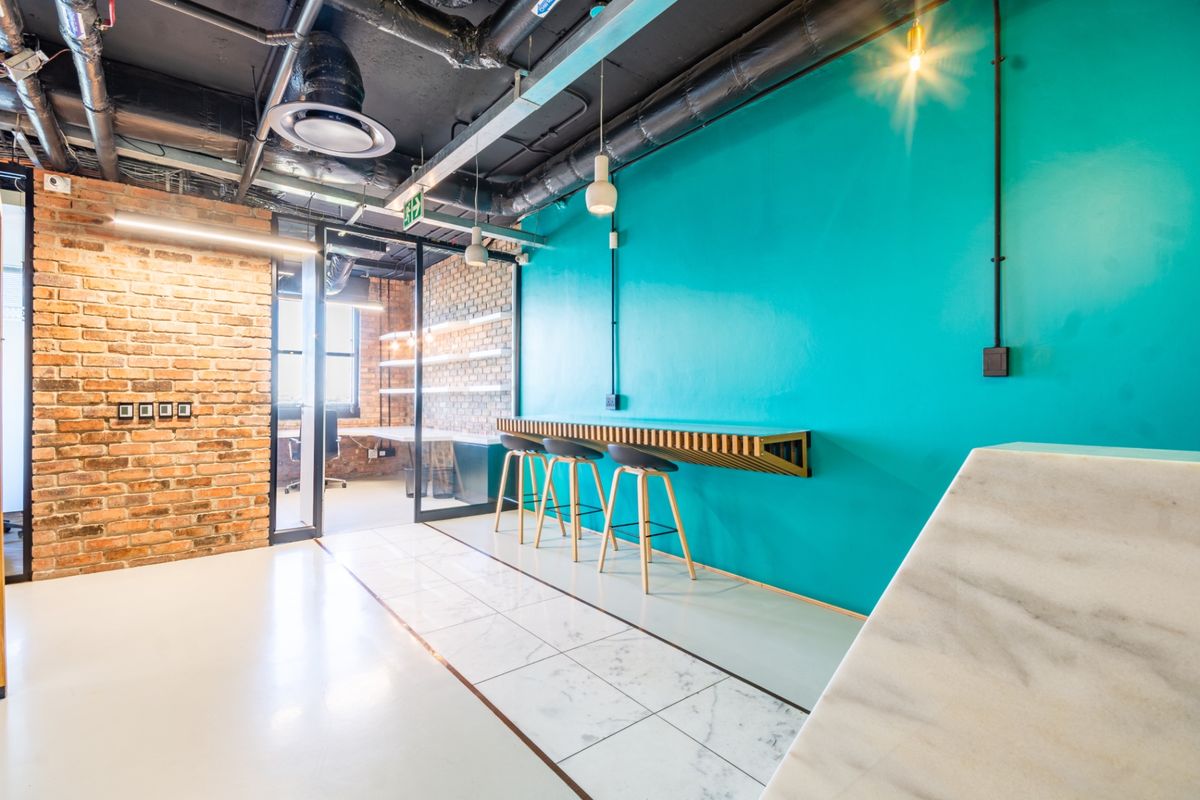 Modern industrial-style office space featuring exposed brickwork, high ceilings with exposed services, glass partitioning, and a sleek built-in wall-mounted bar counter.