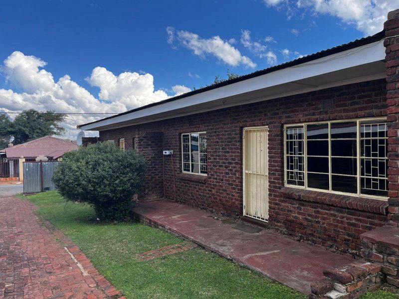 A single-story face brick apartment exterior featuring a paved walkway, security gate, and metal-framed windows under a corrugated metal roof.