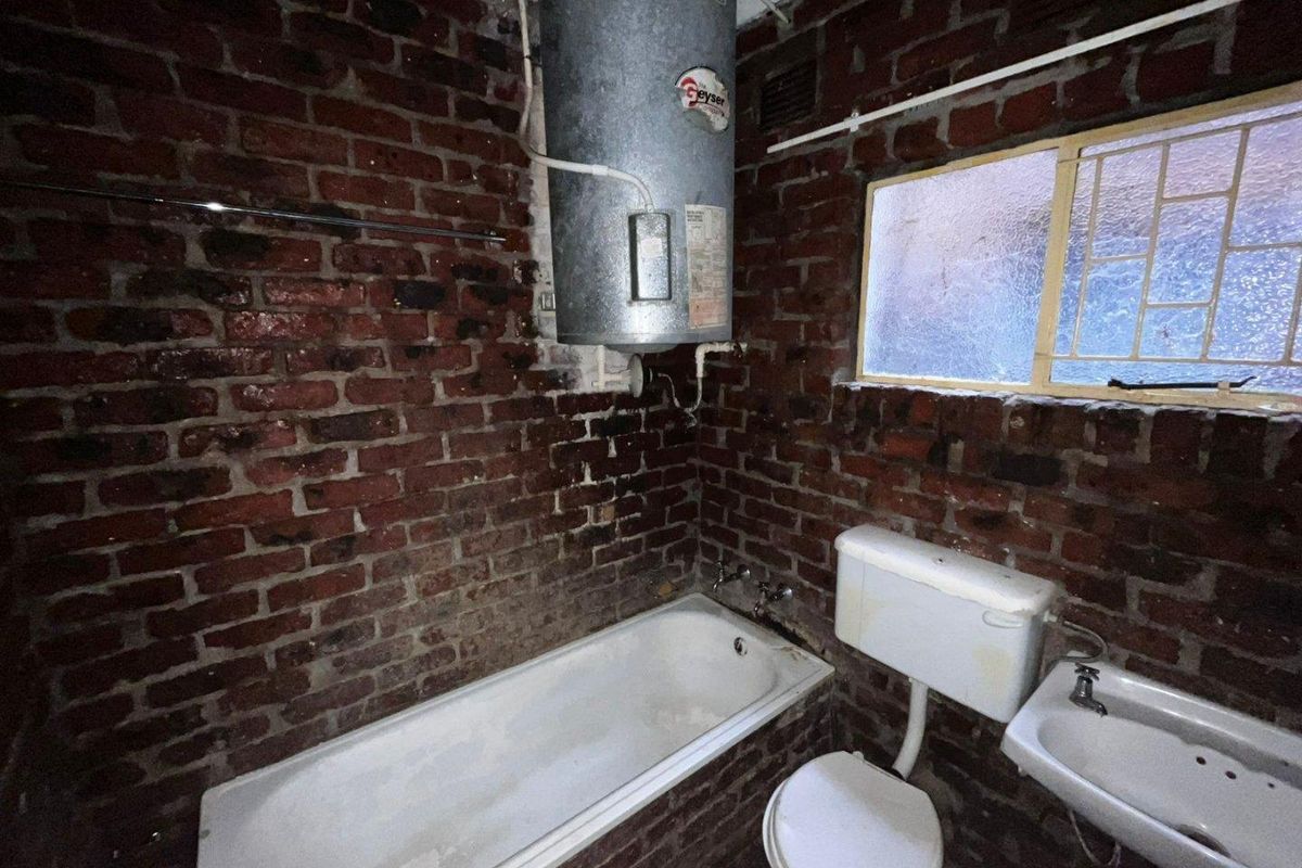 A dated bathroom featuring exposed brick walls, a wall-mounted geyser, a standard bathtub, toilet, and basin, requiring significant renovation and modernization.