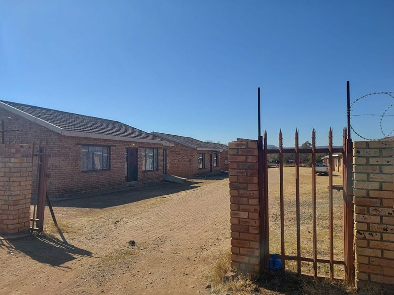 Front view of a face brick townhouse complex featuring a paved driveway, secure metal gate entrance, and perimeter walling with security fencing.
