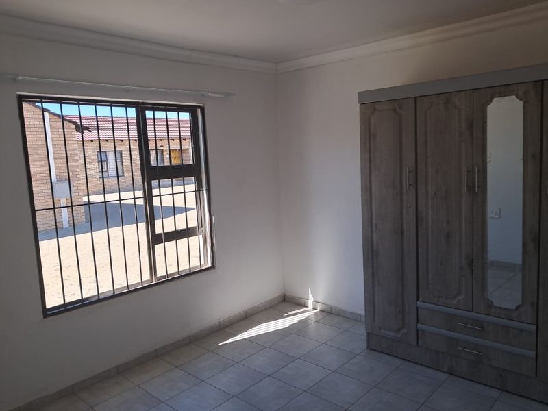 A bedroom featuring tiled flooring, white walls, and a window fitted with security bars, offering a functional and clean living space.