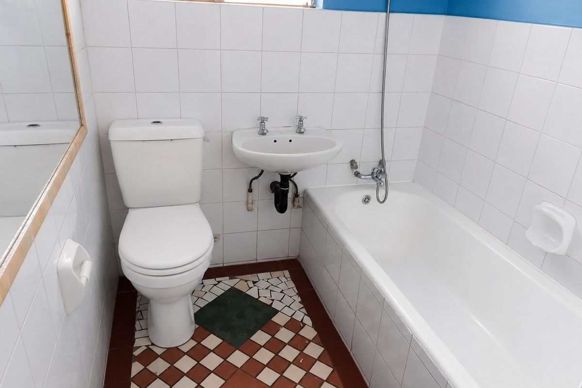 A compact bathroom featuring a white toilet, wall-mounted basin, and bathtub with tiled finishes. The floor requires repair due to missing and damaged tiles.