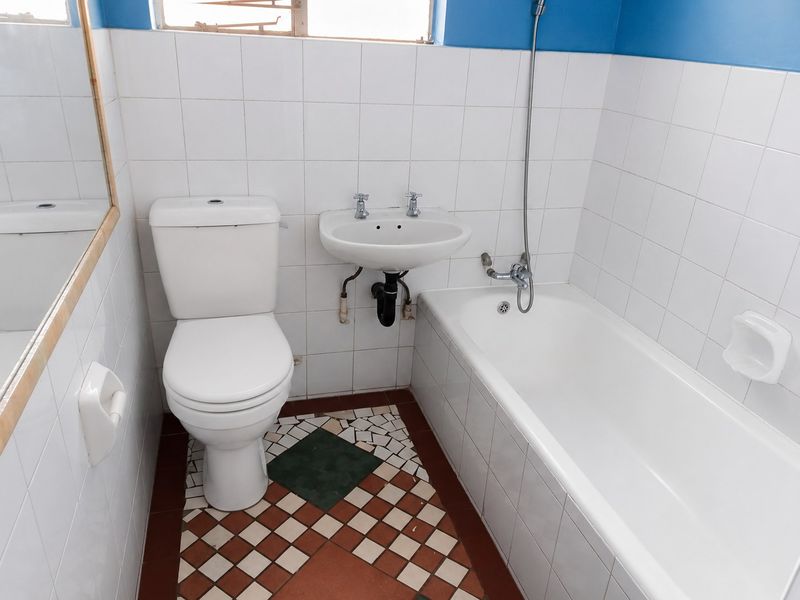 A compact bathroom featuring a white toilet, wall-mounted basin, and bathtub with tiled finishes. The floor requires repair due to missing and damaged tiles.