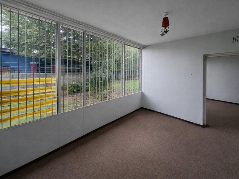 An empty living area featuring carpet flooring, large windows with security bars, and a ventilation grille, offering a blank canvas for renovation.