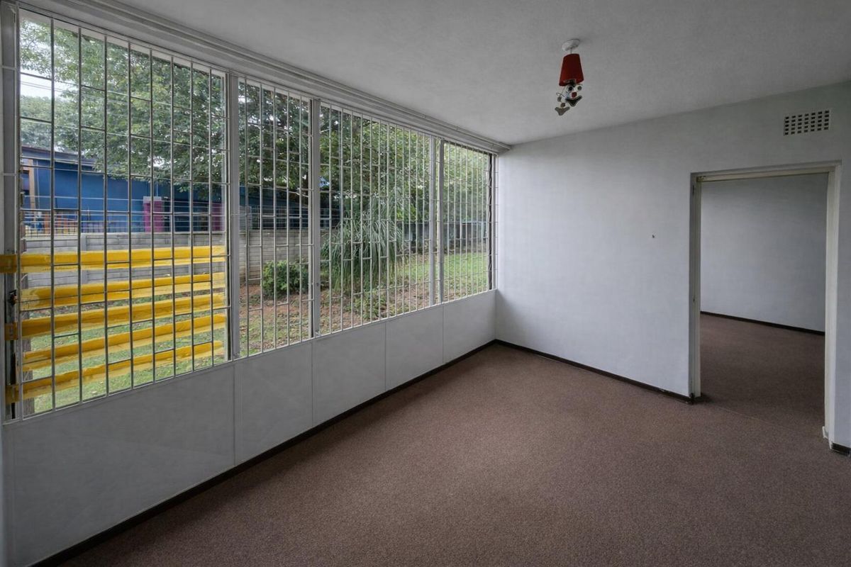 An empty living area featuring carpet flooring, large windows with security bars, and a ventilation grille, offering a blank canvas for renovation.