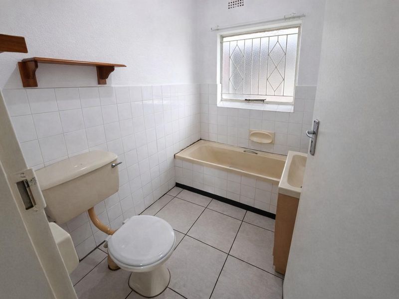 A dated bathroom featuring a bathtub, toilet, and vanity basin with tiled finishes and a security window grille.