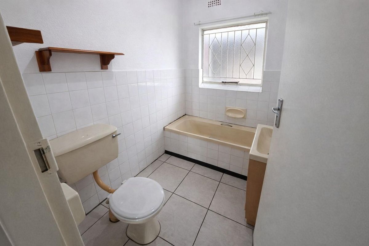 A dated bathroom featuring a bathtub, toilet, and vanity basin with tiled finishes and a security window grille.