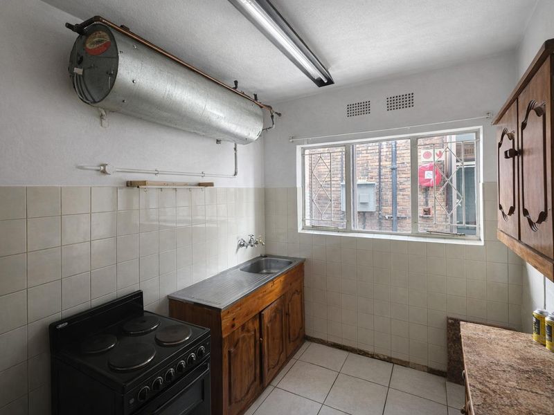 A functional kitchen featuring tiled flooring, a stainless steel sink, wooden cabinetry, and an electric stove, with an exposed geyser unit.