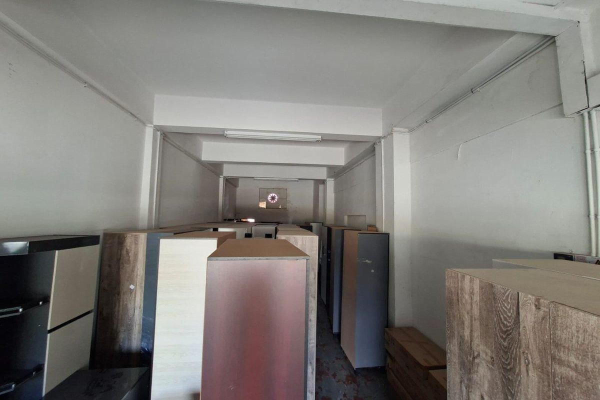 A utilitarian commercial storage or workspace featuring exposed structural beams, functional lighting, and a ventilation fan, currently used for inventory storage.