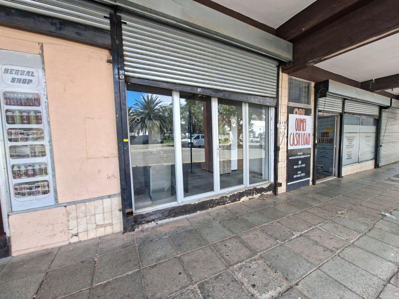 Commercial storefront featuring a glass entrance, metal roller shutter, and tiled pavement access in a street-level retail location.