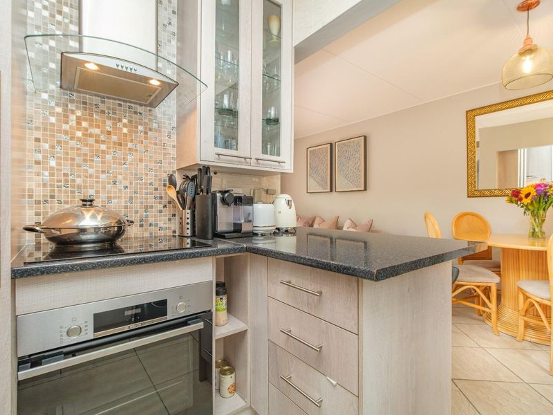 A compact, well-maintained kitchen featuring modern cabinetry, a built-in oven, glass-fronted display cupboards, and a mosaic tile backsplash, seamlessly flowing into the dining area.