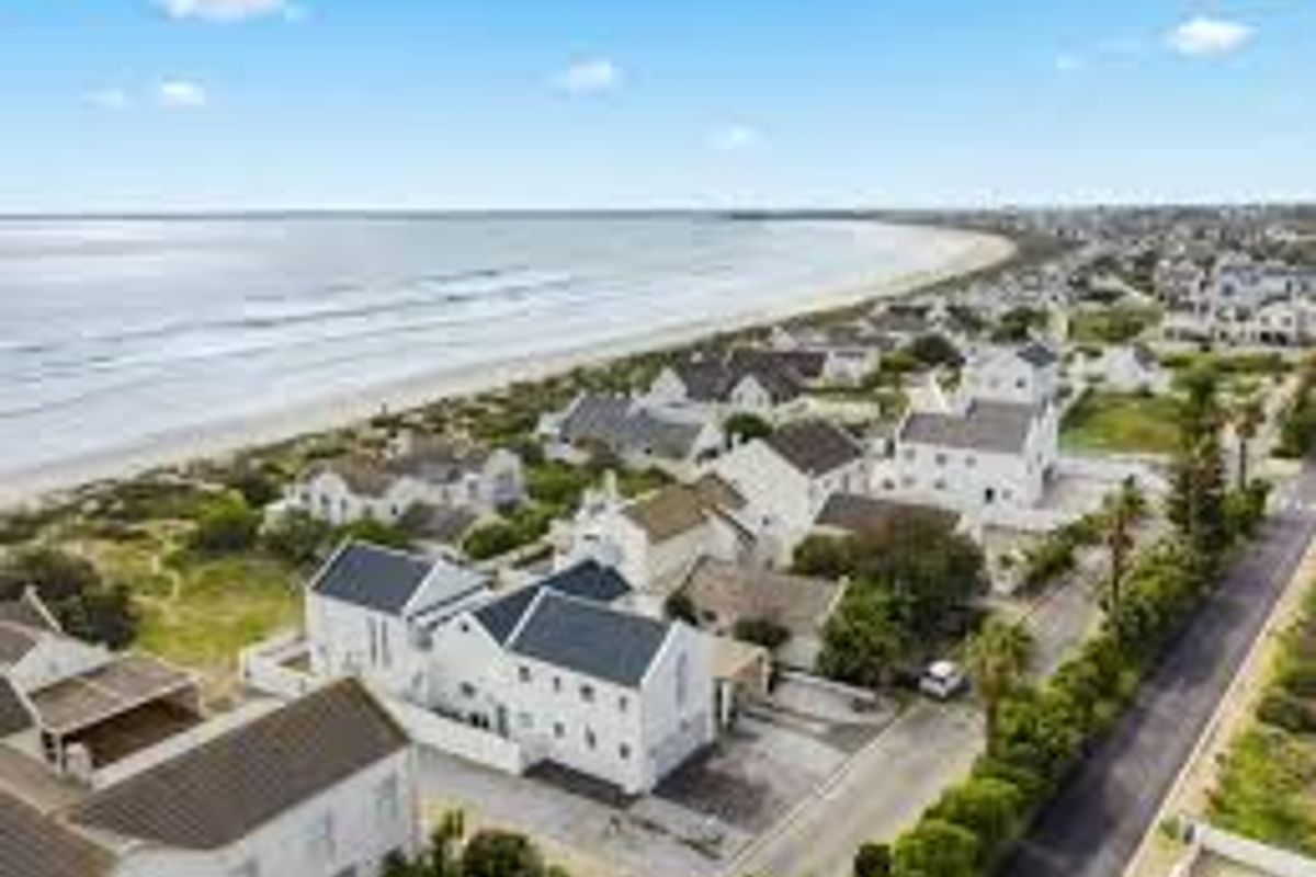 An aerial view of a coastal residential estate featuring white houses with dark pitched roofs, situated directly adjacent to the beach and ocean.