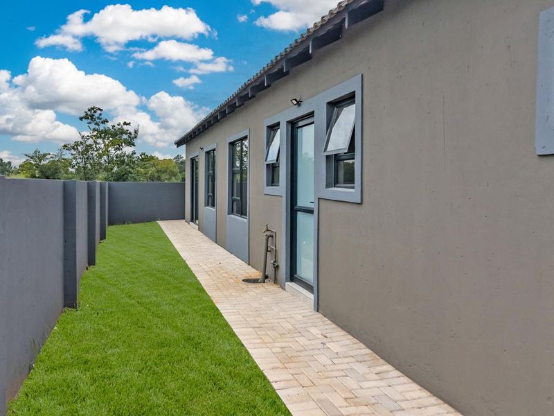 A well-maintained rear exterior view featuring a paved walkway, manicured lawn, and a solid boundary wall along the side of the house.
