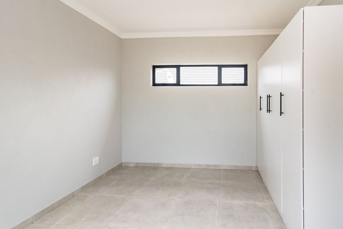 A clean, modern bedroom featuring neutral tiled flooring, built-in white cupboards with black hardware, and a horizontal clerestory window.