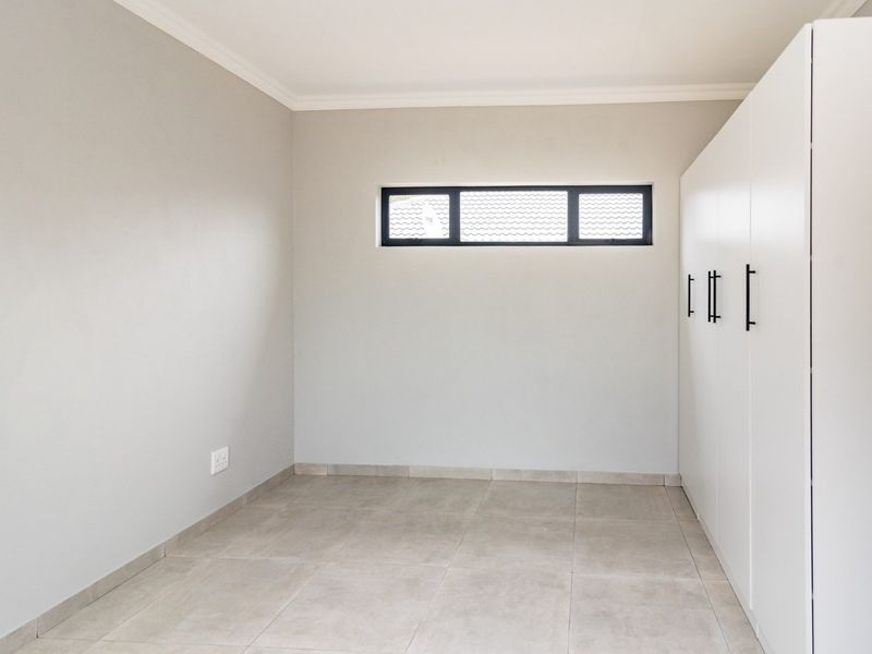 A clean, modern bedroom featuring neutral tiled flooring, built-in white cupboards with black hardware, and a horizontal clerestory window.