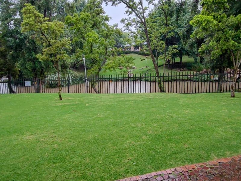 A well-maintained garden area featuring a lush green lawn, paved pathway, and secure metal perimeter fencing overlooking a natural pond and mature trees.