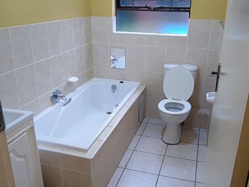 A functional bathroom featuring a built-in bathtub, toilet, and vanity cabinet with neutral wall and floor tiling.
