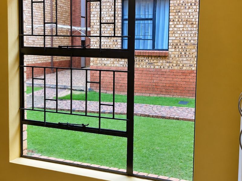 A bright bedroom featuring yellow walls, carpeted flooring, and a large window with security bars overlooking a communal garden area.
