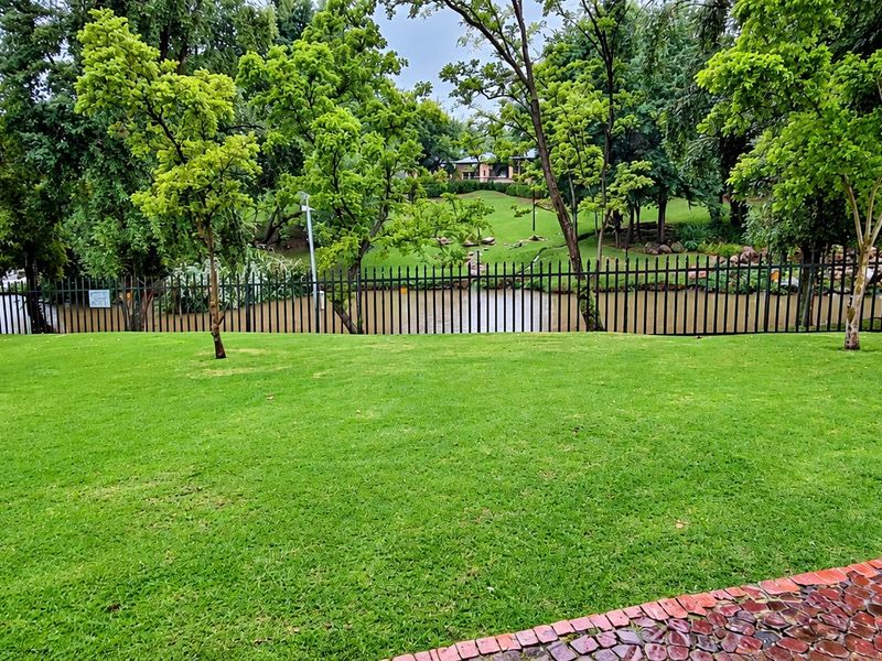 A well-maintained garden area featuring a lush green lawn, a brick paved pathway, and secure metal perimeter fencing overlooking a scenic water feature.
