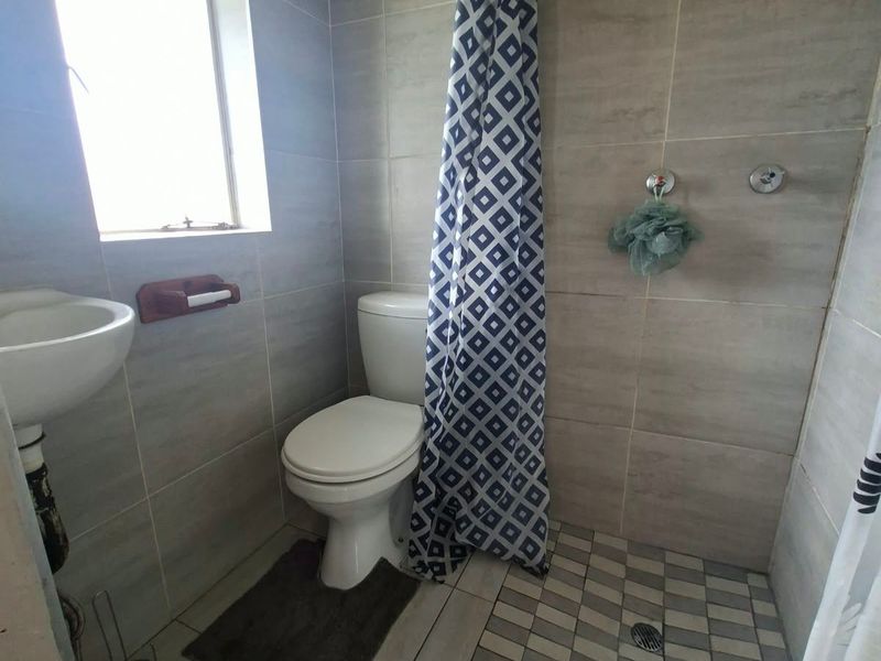 A compact bathroom featuring tiled walls and flooring, a wall-mounted basin, a toilet, and a shower area with a fixed shower head.