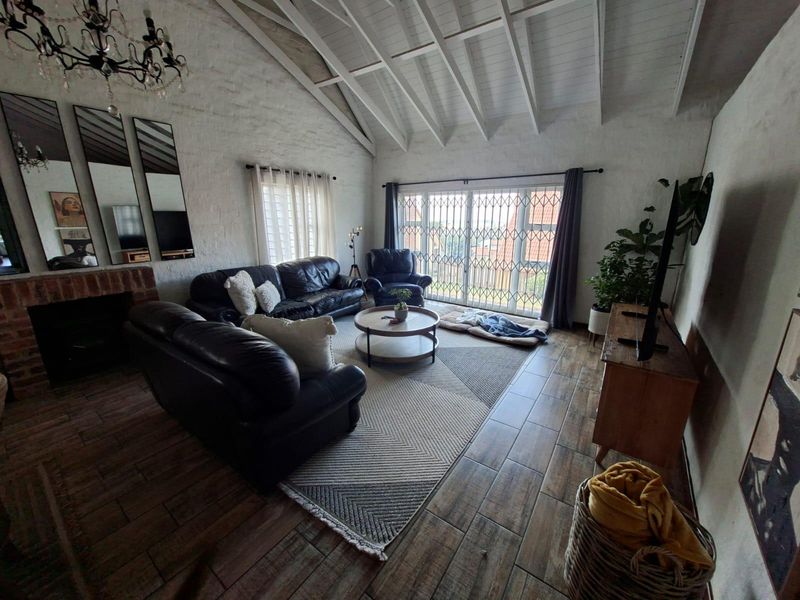 Spacious living room featuring a vaulted ceiling with exposed beams, tiled flooring, and a brick fireplace, opening to an outdoor area through sliding doors.