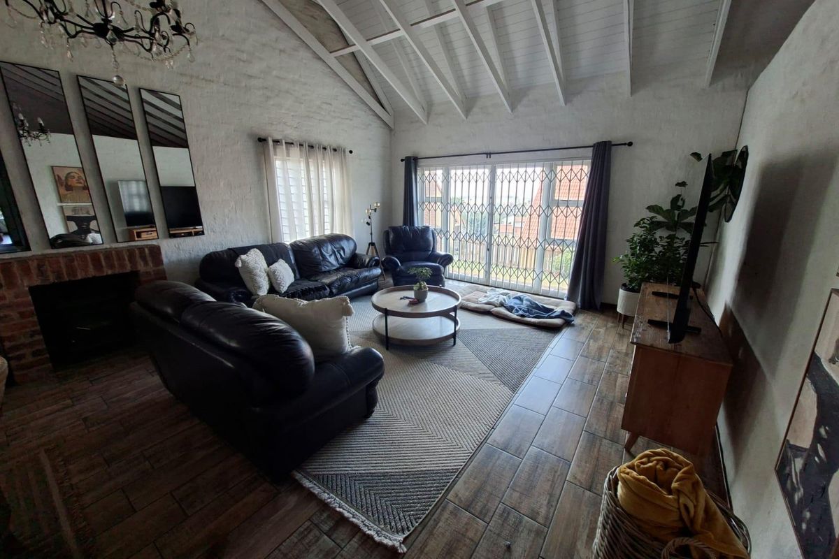 Spacious living room featuring a vaulted ceiling with exposed beams, tiled flooring, and a brick fireplace, opening to an outdoor area through sliding doors.