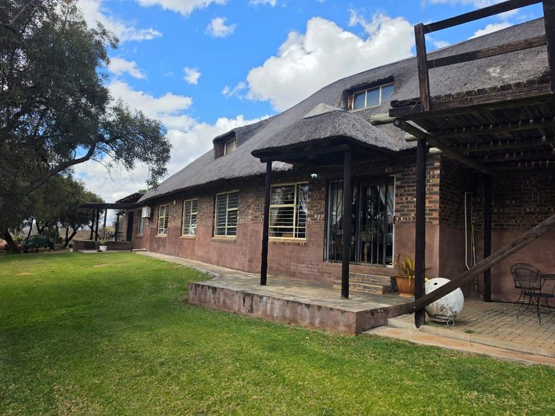 Rear view of a traditional thatch-roof house featuring a covered patio, brick facade, and an elevated timber balcony overlooking a spacious garden.