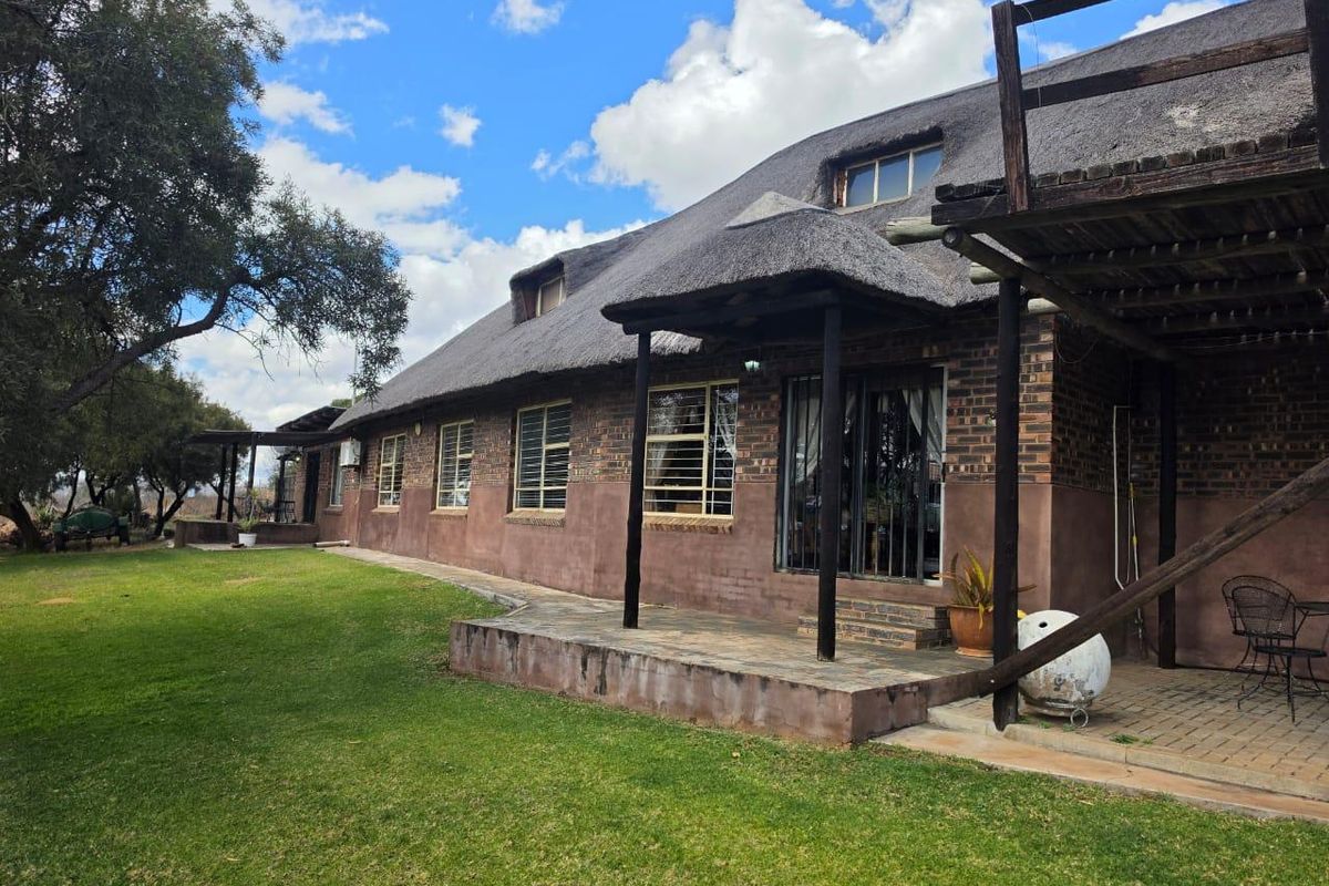 Rear view of a traditional thatch-roof house featuring a covered patio, brick facade, and an elevated timber balcony overlooking a spacious garden.