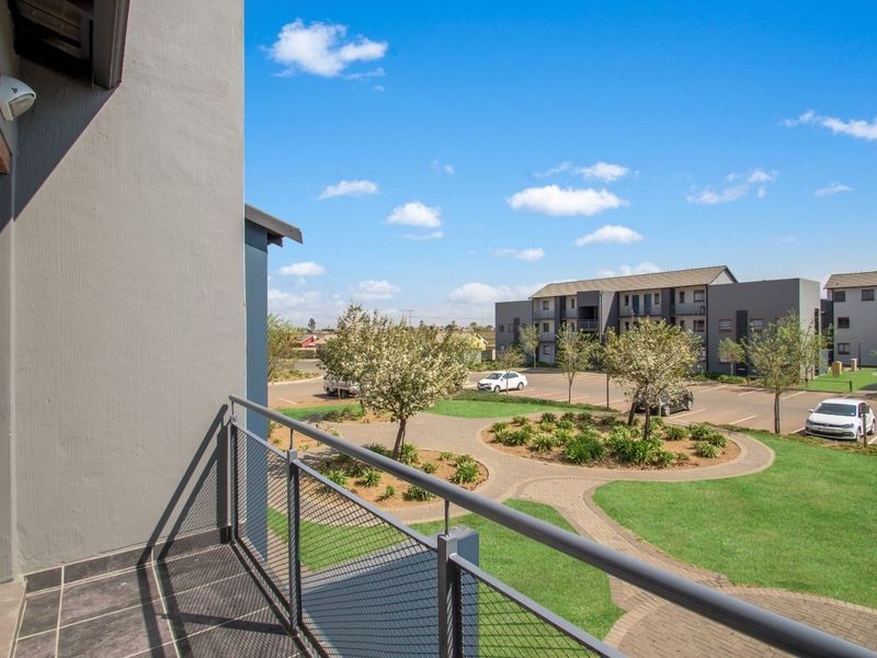 A private balcony with tiled flooring and a metal balustrade, overlooking the apartment complex grounds and parking area.