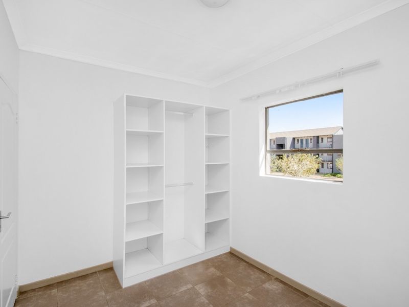 A bright, empty bedroom featuring tiled flooring, a built-in white shelving unit, and a window with a view of the surrounding apartment complex.